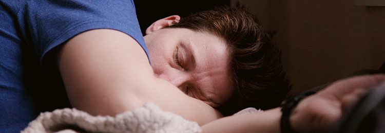 A man in a purple shirt sleeping on his side with his arm tucked under the pillow.